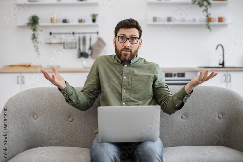 Confused bearded male wearing eyeglasses and casual attire shrugging shoulders while sitting on couch with modern laptop and looking at camera. Concept of doubt, people and technology.