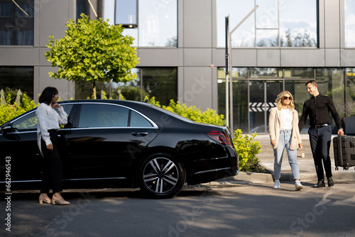 Man helps a businesswoman to carry her suitcase into a luxury car from an office building or hotel. Female chauffeur waits near a car. Concept of business trips, and luxury transfer services