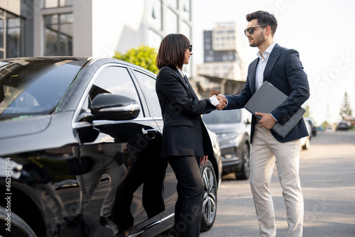 Business man and business woman shaking hands with each other  standing in front of the car outdoors. Concept of successful deal and partnership