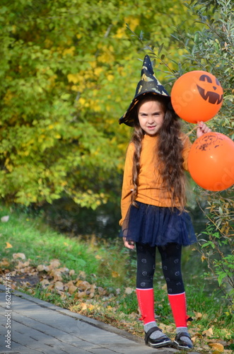 happy laughing child girl in witch costume to halloween. with a jack-o -lantern candy bowl  trick or treat  in a witch s hat  with colorful balls in the autumn forest  eating spider-shaped candy