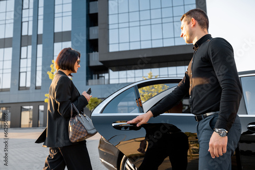 Chauffeur helps an elegant business woman gets in car, opening door of a premium taxi near office building or hotel. Concept of personal driver or business trips photo