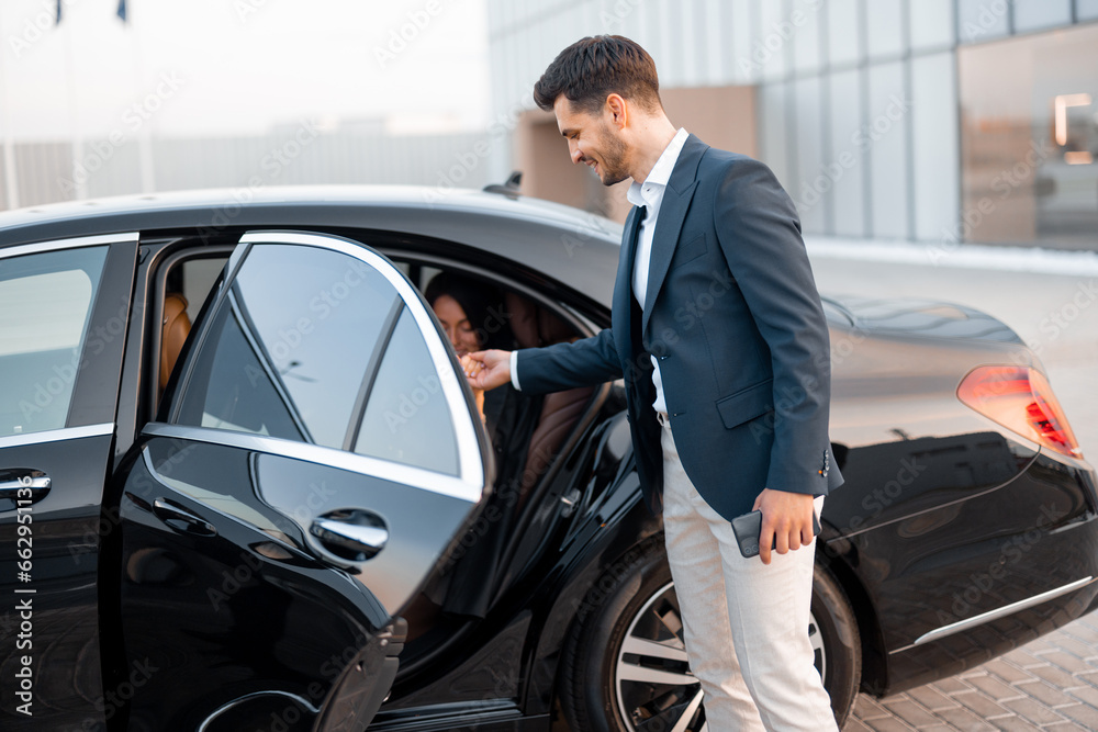 Businessman helps gently a woman to get out of a car, arrived by luxury vehicle for some event at evening. Concept of transportation and business lifestyle