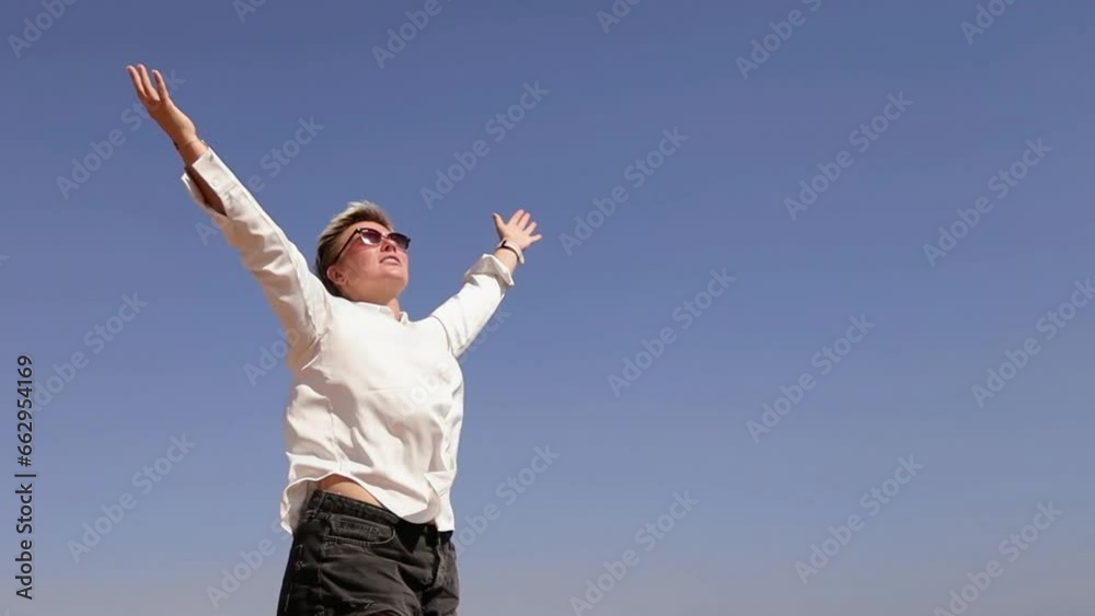 Happy smiling blond short haired woman raising arms against blue sky on sunny summer day Happy smiling blond short haired woman raising arms against blue sky on sunny summer day