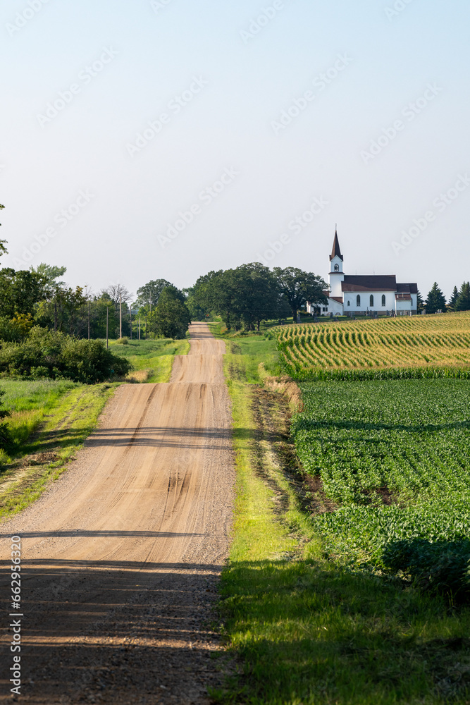 Fototapeta premium Looking down a country road lined with fields and a church on a sunny day in rural Minnesota, USA. 