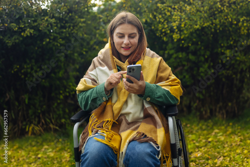 Young woman in bright stylish clothes with physical disability uses a mobile phone, reads news, communicates on social networks sitting in a wheelchair in park in autumn nature. High quality photo © Shi 
