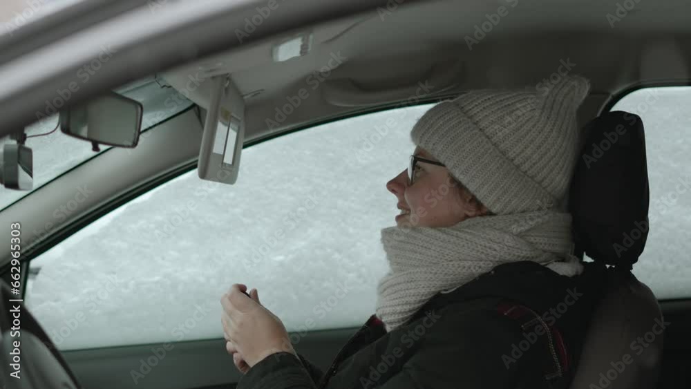 European woman flips back sun visor with mirror and paints her lips in snowy car. Quick makeup in warm clothes while waiting for driver in snowfall. Young single woman preparing for date. European woman flips back sun visor with mirror and paints her lips in snowy car. Quick makeup in warm clothes while waiting for driver in snowfall. Young single woman preparing for date.