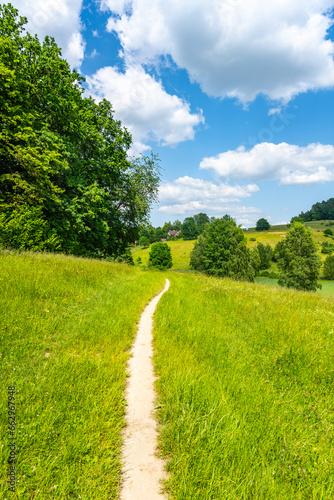 Fotografie Narrow rural pathway in hilly landscape with lush green meadows on sunny summer day