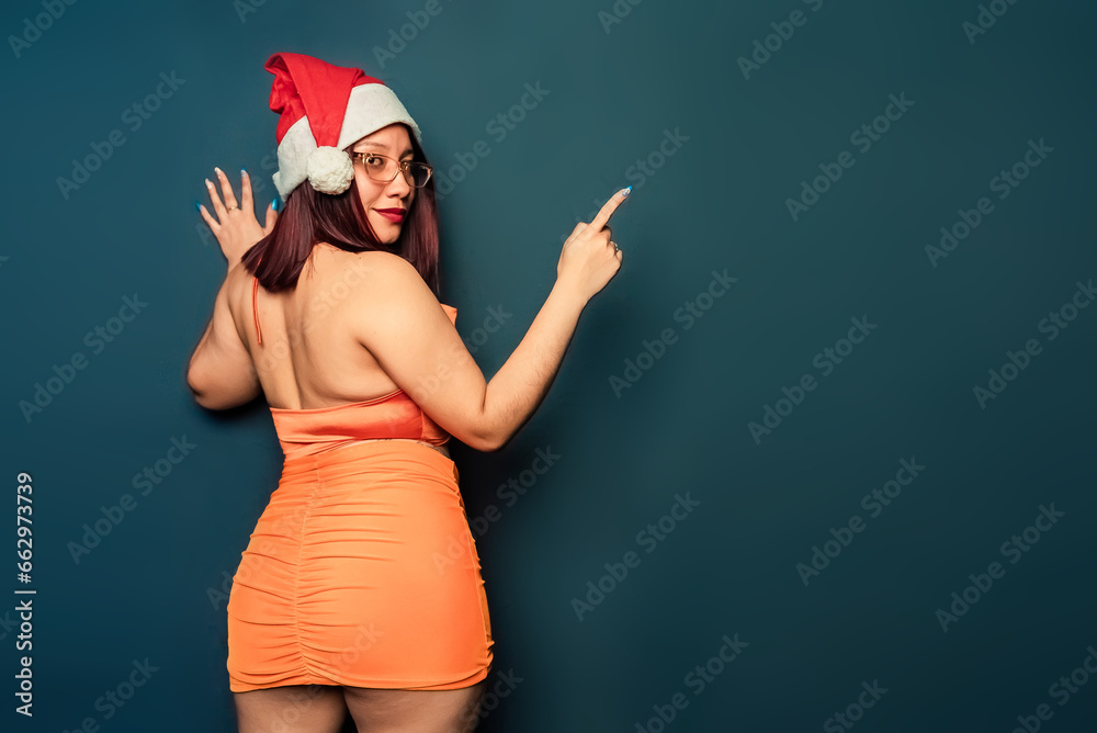 Beautiful woman at Christmas time wearing a red and white Christmas hat and an orange dress pointing with her finger. Christmas concept. santa claus concept. copy space. Beautiful woman at Christmas time wearing a red and white Christmas hat and an orange dress pointing with her finger. Christmas concept. santa claus concept. copy space.