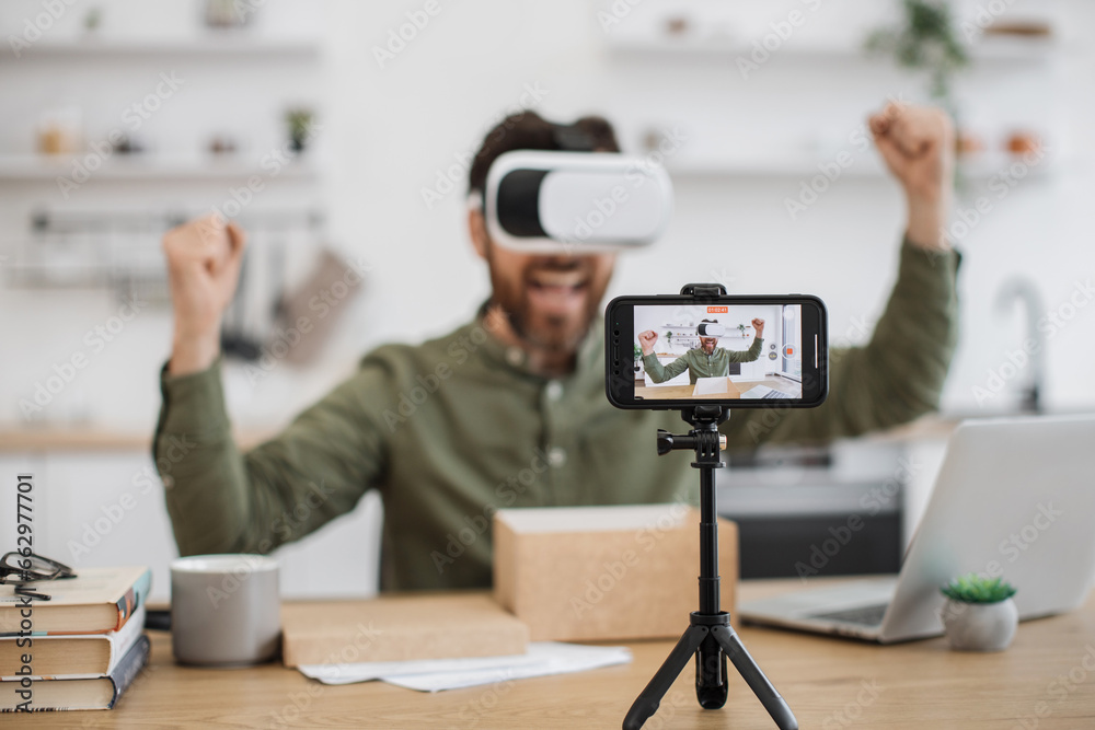 Modern smartphone recording video of happy male blogger testing new VR headset and making review for subscribers on social media. Innovative electronics and online purchase concept. Modern smartphone recording video of happy male blogger testing new VR headset and making review for subscribers on social media. Innovative electronics and online purchase concept.