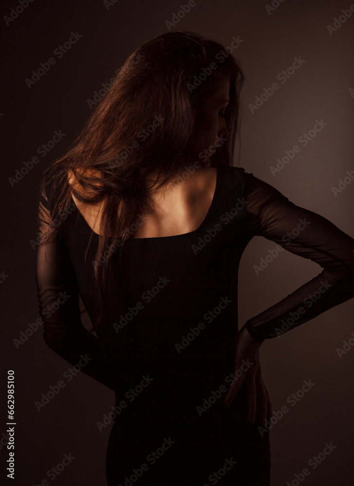 Beautiful sexy slim elegant woman standing with healthy long hair in style fashion black dress on black shadow dark background with empty copy space. Closeup back view portrait
