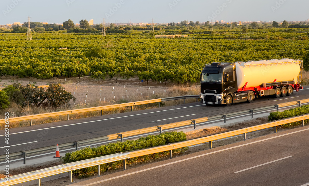 Isothermal Tank truck driving on highway at farm field. Oil and Gas Transportation Logistics. Cistern tanker with petrochemicals. Liquid Chemical Freight and foodstuff. Truck on road on sunset.