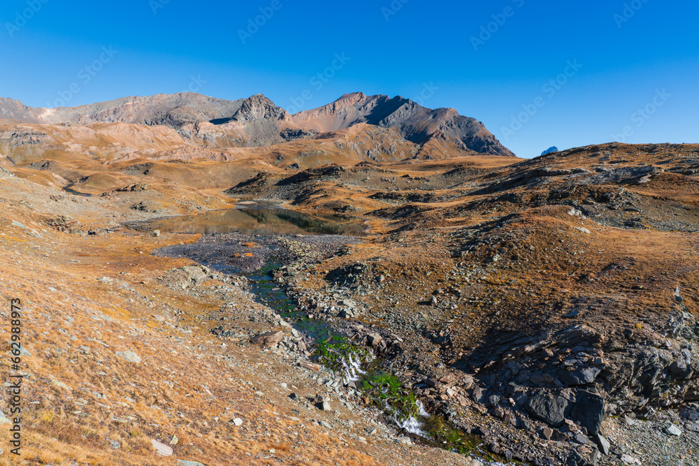 Mountain landscape in autumn, blue sky with no clouds, quiet lake with reflection and water stream with green algae