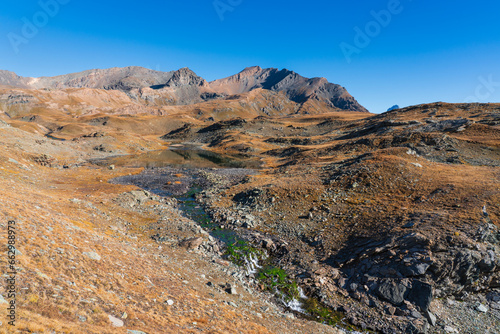 Mountain landscape in autumn, blue sky with no clouds, quiet lake with reflection and water stream with green algae