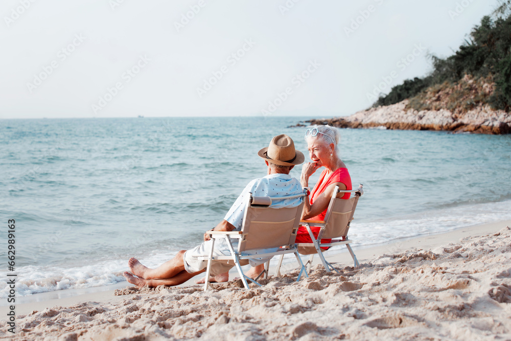 Beautiful and happy senior couple in love sitting on the beach at sunset and enjoying amazing sea view at tropical sunset together