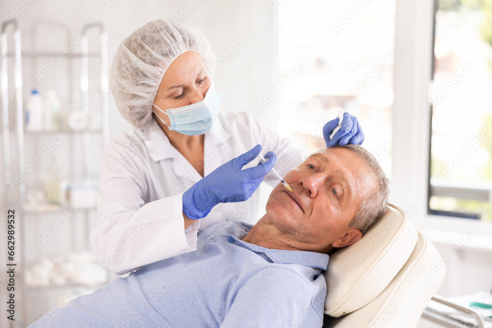 Obraz premium Experienced female cosmetologist working with senior male client in clinic of aesthetic medicine, performing minimally invasive procedure using filler injections to correct lips contour