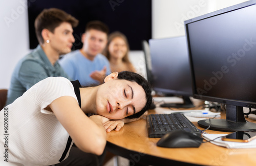 Exhausted young girl sleeping at table in classroom with computers and fellow students due to lengthy educational session or uninteresting lesson topic