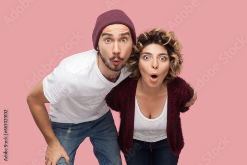 Portrait of attractive surprised shocked couple of friends standing together looking at camera very attentive, expressing shock and astonishment. Indoor studio shot isolated on pink background.