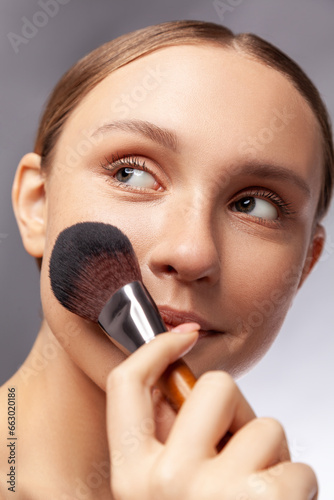Closeup portrait of beautiful woman holding makeup brush applying powder looking smiling away beauty visage procedures. Indoor studio shot isolated over gray background.