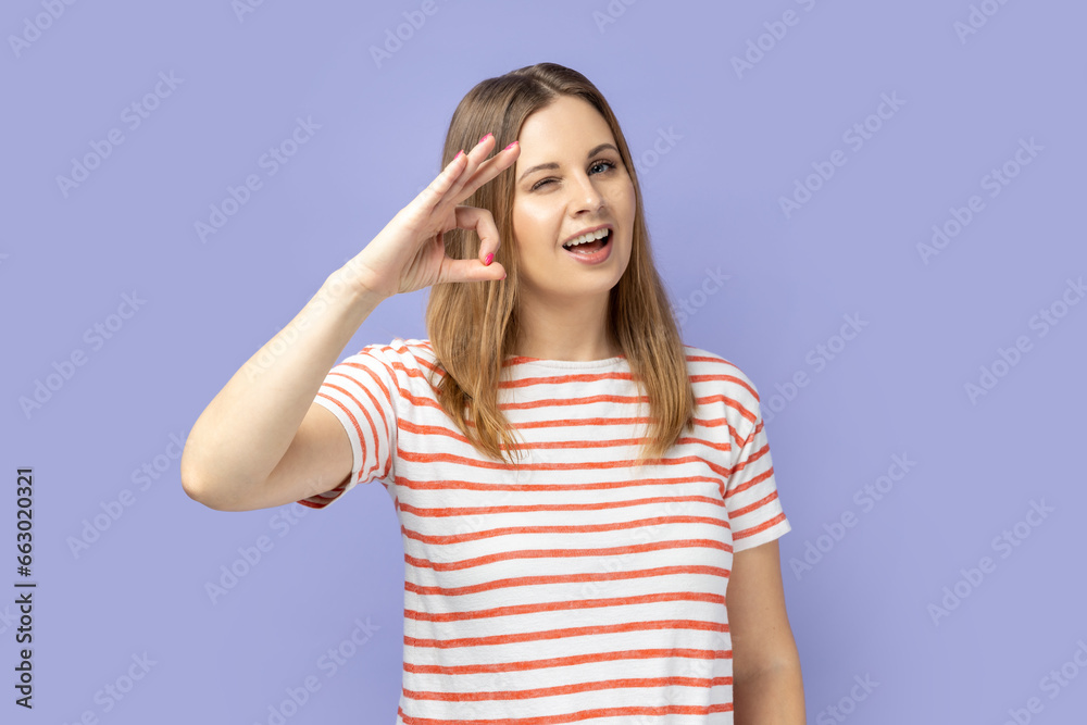 Portrait of cheerful young adult blond woman wearing striped T-shirt showing okay sign, assures you everything is fine, looks gladly. Indoor studio shot isolated on purple background. Portrait of cheerful young adult blond woman wearing striped T-shirt showing okay sign, assures you everything is fine, looks gladly. Indoor studio shot isolated on purple background.