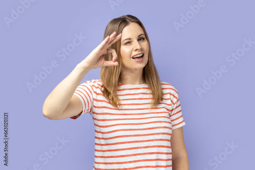 Portrait of cheerful young adult blond woman wearing striped T-shirt showing okay sign, assures you everything is fine, looks gladly. Indoor studio shot isolated on purple background.