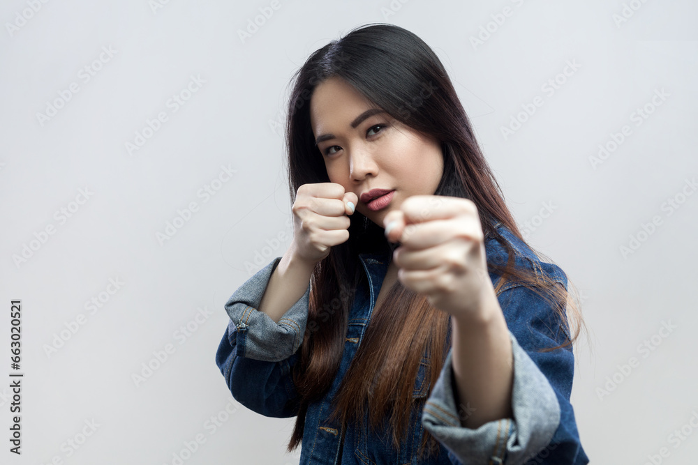 Portrait of angry aggressive beautiful serious brunette woman in blue denim jacket standing with clenched fists, fighting with somebody. Indoor studio shot isolated on gray background. Portrait of angry aggressive beautiful serious brunette woman in blue denim jacket standing with clenched fists, fighting with somebody. Indoor studio shot isolated on gray background.