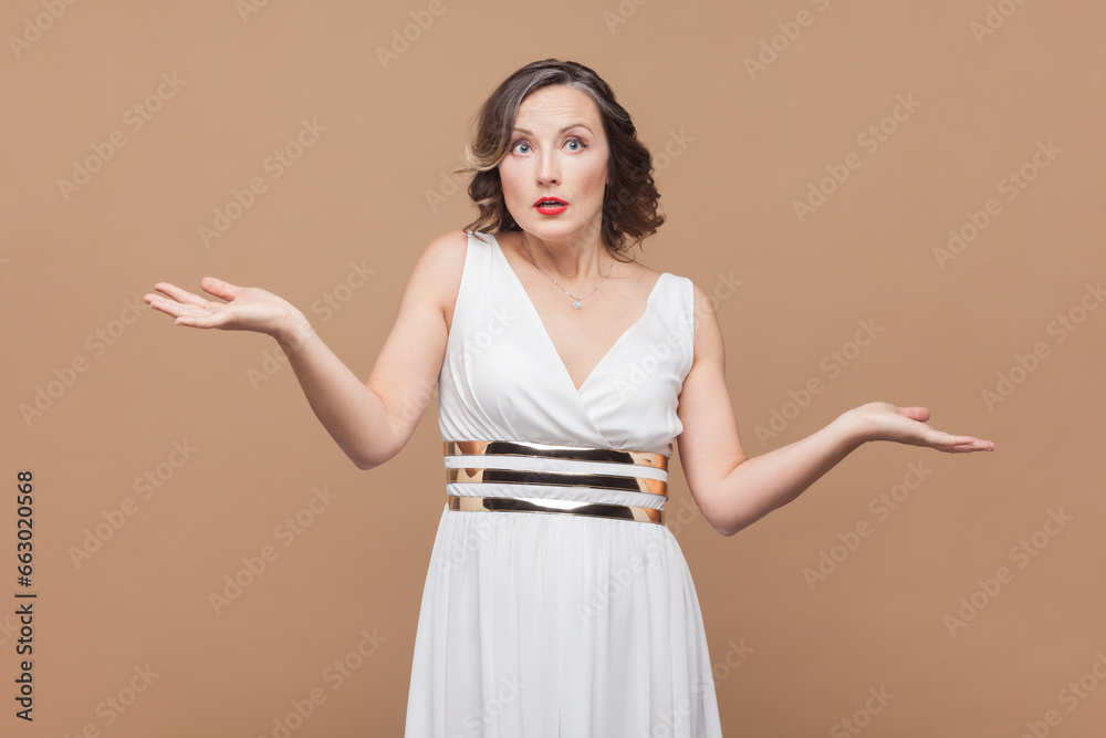 I do not know. Uncertain woman shruging shoulders, keeps palms sideways, has questioned perplexed face expression, no idea what to do. Indoor studio shot isolated on light brown background. I do not know. Uncertain woman shruging shoulders, keeps palms sideways, has questioned perplexed face expression, no idea what to do. Indoor studio shot isolated on light brown background.