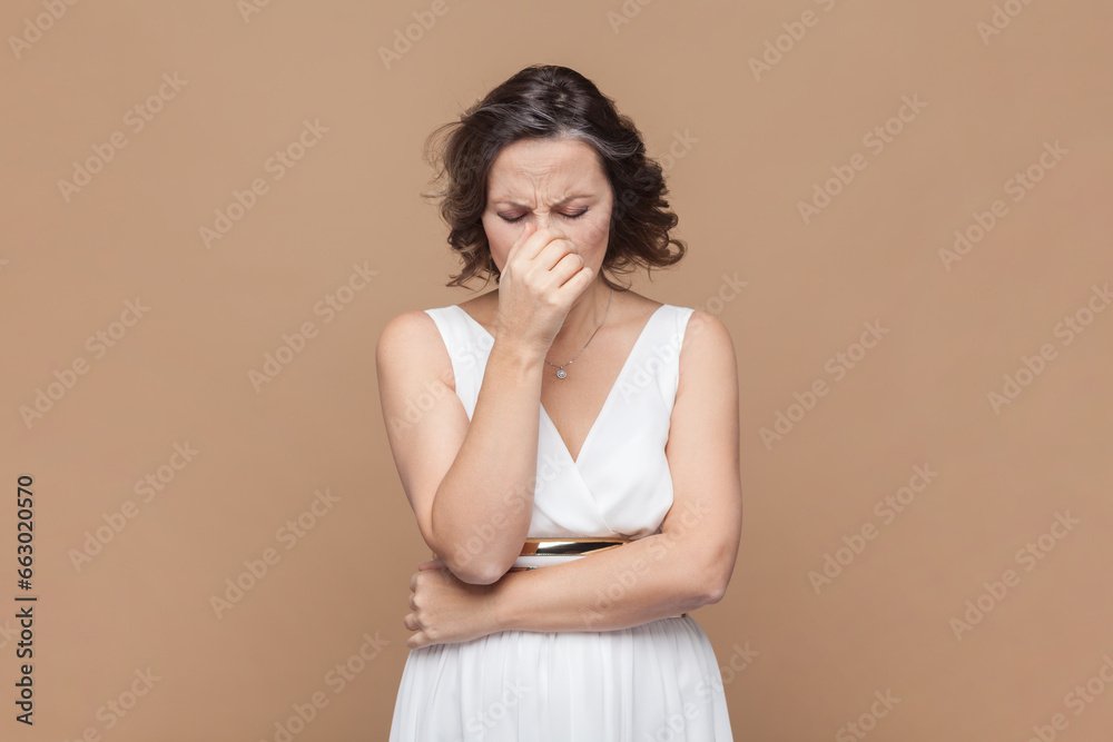Portrait of sad crying disappointed middle aged woman with wavy hair looking unhappy crying, having problems, wearing white dress. Indoor studio shot isolated on light brown background. Portrait of sad crying disappointed middle aged woman with wavy hair looking unhappy crying, having problems, wearing white dress. Indoor studio shot isolated on light brown background.