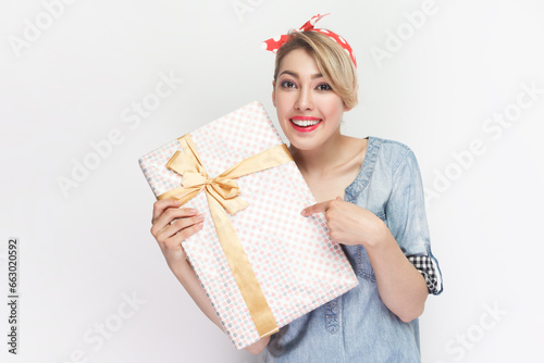 Portrait of charming smiling satisfied blonde woman wearing blue denim shirt and red headband standing pointing at gift box, present for holiday. Indoor studio shot isolated on gray background.