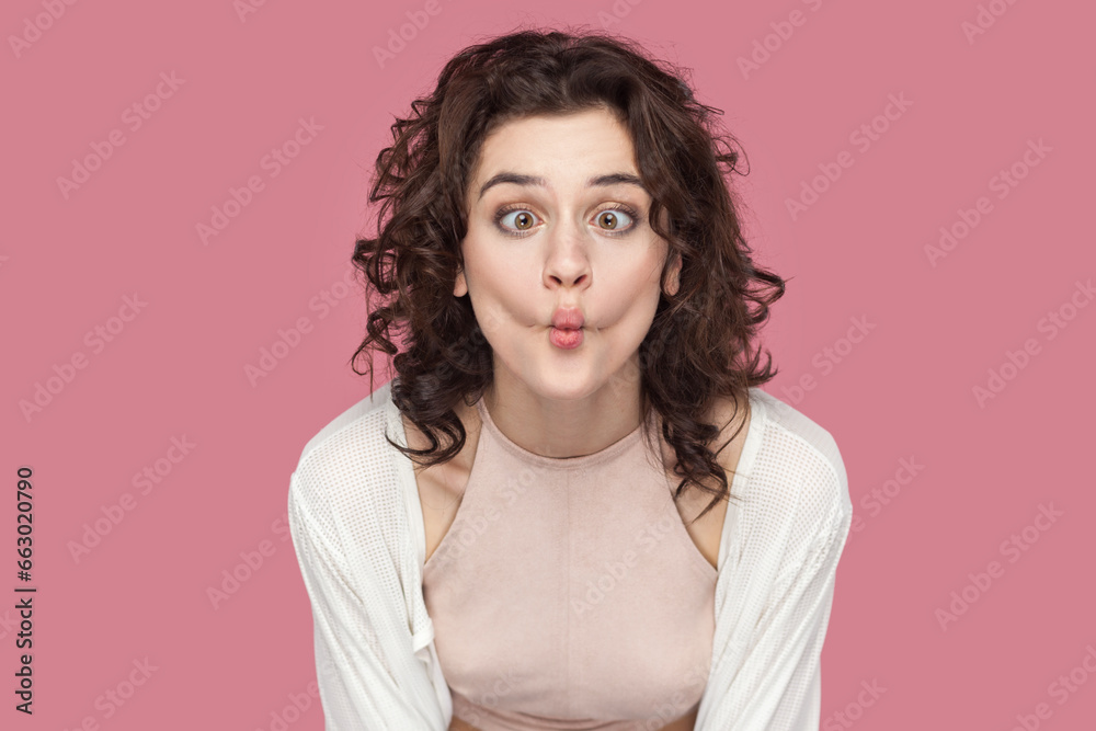 Fototapeta premium Portrait of funny positive woman with curly hair wearing casual style outfit having fun, making fish, demonstrates childish behavior. Indoor studio shot isolated on pink background.