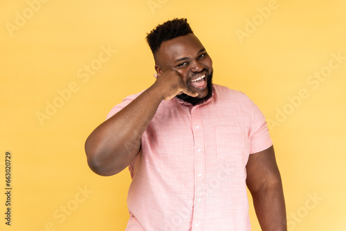 Portrait of satisfied man wearing pink shirt flirting holding fingers near ear showing call gesture with toothy smile, answering call. Indoor studio shot isolated on yellow background.