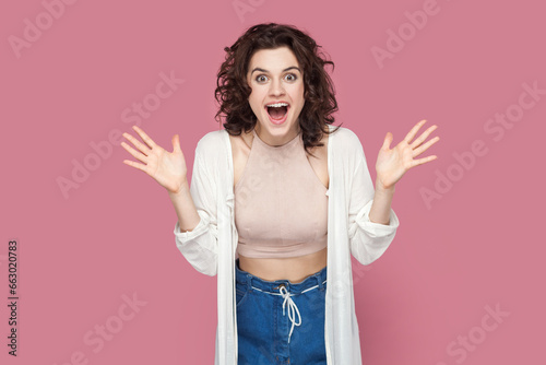 Portrait of positive smiling excited woman with curly hair wearing casual style outfit, raised her arms, sees something excitement. Indoor studio shot isolated on pink background.