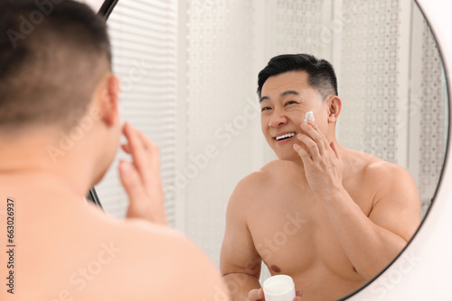 Handsome man applying cream onto his face near mirror indoors