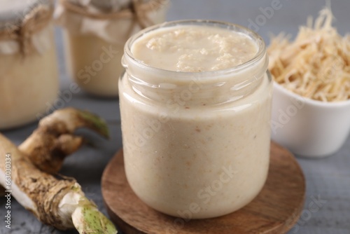 Spicy horseradish sauce in jar and roots on table, closeup photo