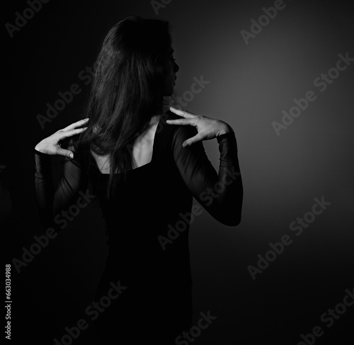 Beautiful slim elegant woman standing with long hair in fashion black dress on shadow dark background holding hands on the shoulders with empty copy space. Closeup back view portrait. Art
