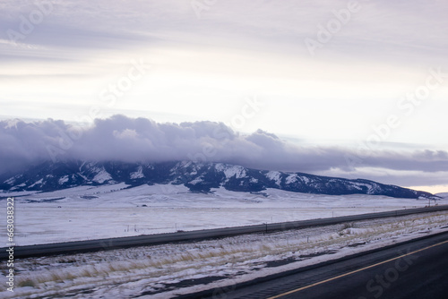 Beautiful winter landscape with a highway between blue snowy mountains, fluffy clouds on a sunset orange sky. Winter snow background. Elk Mountain, Wyoming, USA. Picturesque winter sky
