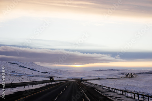 Beautiful winter landscape with a highway between blue snowy mountains, fluffy clouds on a sunset orange sky. Winter snow background. Elk Mountain, Wyoming, USA. Picturesque winter sky