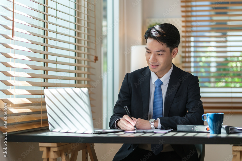 Focused asian businessman writing important information on notepad and using laptop at working desk. Focused asian businessman writing important information on notepad and using laptop at working desk.