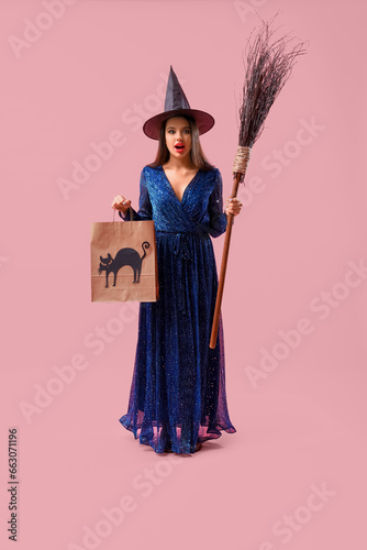 Surprised young woman dressed for Halloween as witch with broom and gift bag on pink background