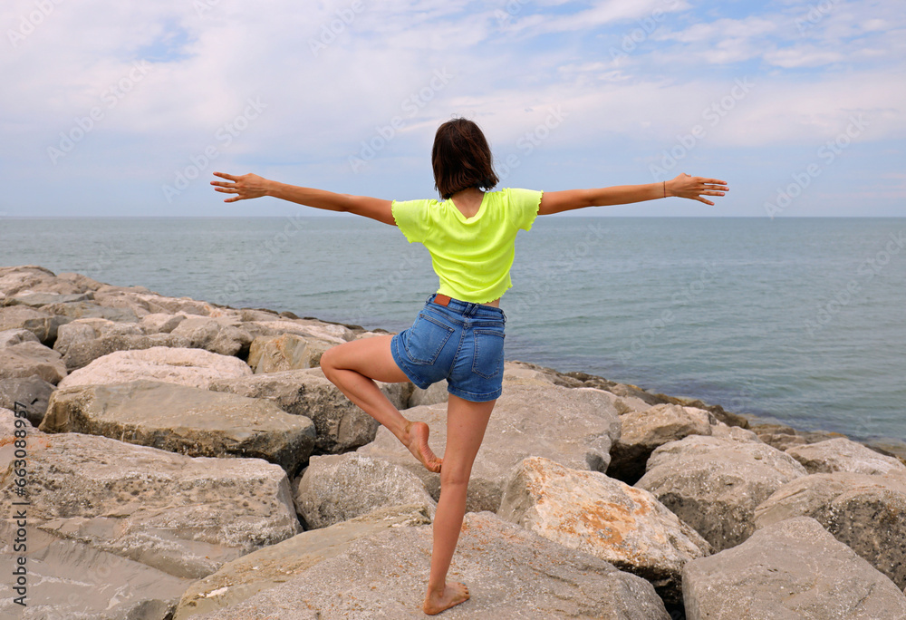 young slender girl performs gymnastic exercises on the rocks by the sea in jeans shorts young slender girl performs gymnastic exercises on the rocks by the sea in jeans shorts