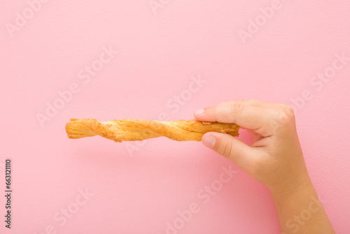Baby girl hand fingers holding and showing bread stick with cheese on light pink table background. Pastel color. Closeup. Salty snack. Top down view.