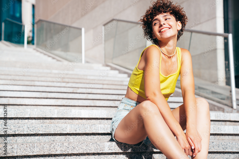 Young beautiful smiling hipster woman in trendy summer clothes. Carefree woman with curls hairstyle, posing in the street at sunny day. Positive model outdoors. Cheerful and happy. Sits at the stairs