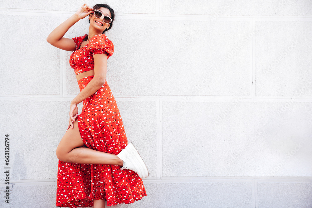 Young beautiful smiling hipster woman in trendy summer red dress. Carefree woman posing in the street near wall. Positive model outdoors in at sunny day. Cheerful and happy. In sunglasses Young beautiful smiling hipster woman in trendy summer red dress. Carefree woman posing in the street near wall. Positive model outdoors in at sunny day. Cheerful and happy. In sunglasses