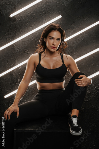 Fit woman in sportswear sits on a black cube, brightly lit lamps illuminating the dark studio