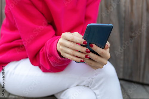 Close up Woman hands in red hoodie with red manicure is holding smartphone on bench outdoors.