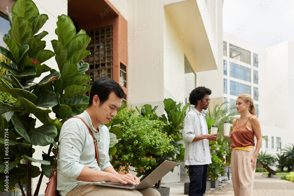 Young man working on laptop at office entrance when his coworkers discussing upcoming meeting Young man working on laptop at office entrance when his coworkers discussing upcoming meeting