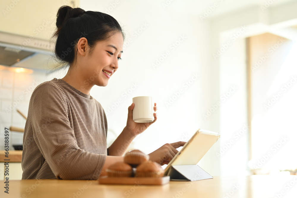 Attractive young woman drinking coffee and using digital tablet at table in kitchen Attractive young woman drinking coffee and using digital tablet at table in kitchen