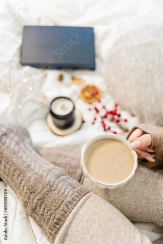 Cozy winter scene. Christmas vibes. Woolen socks. Woman having coffee in bed with book and candle.