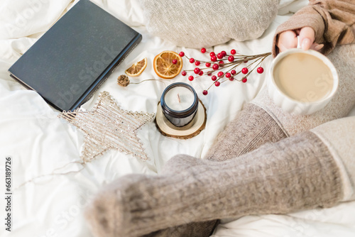 Cozy winter scene. Christmas vibes. Woolen socks. Woman having coffee in bed with book and candle.