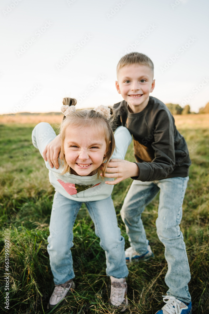 Fototapeta premium Happy children walking spending time together in nature. Daughter, son hold hands and run in green grass in field at sunset. Concept family holiday outdoors. Kids playing in mountains on autumn day.