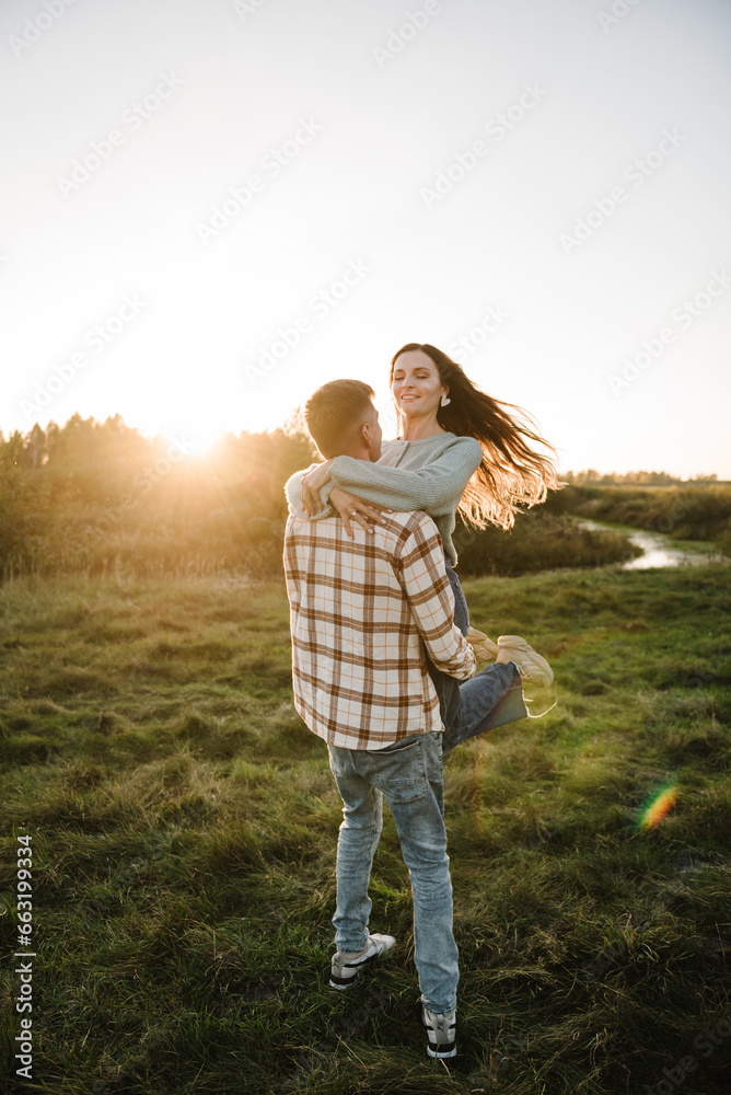 Young man spins woman, walking spending time together in nature. Couple hugging and smiling in grass in field at sunset. Family holiday outdoors. Female embrace male in summer park. Sunset scene.